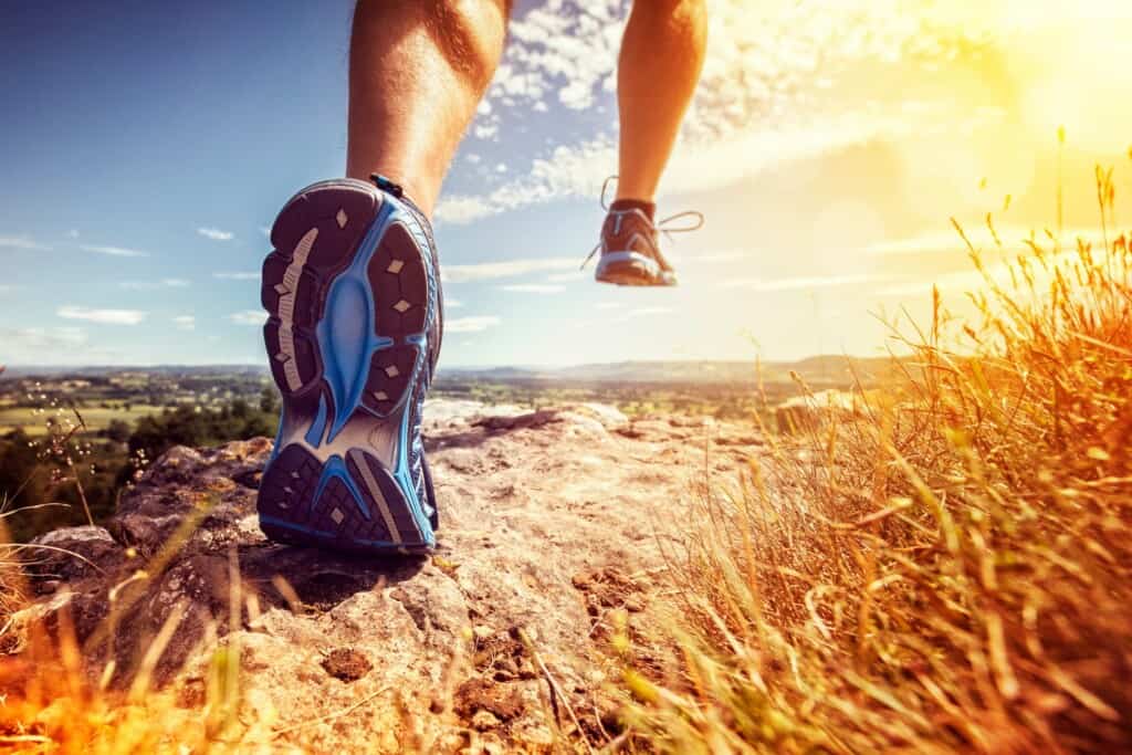 Close-up of a person's legs and running shoes as they run on a rocky terrain with a scenic landscape and bright sunlight in the background.