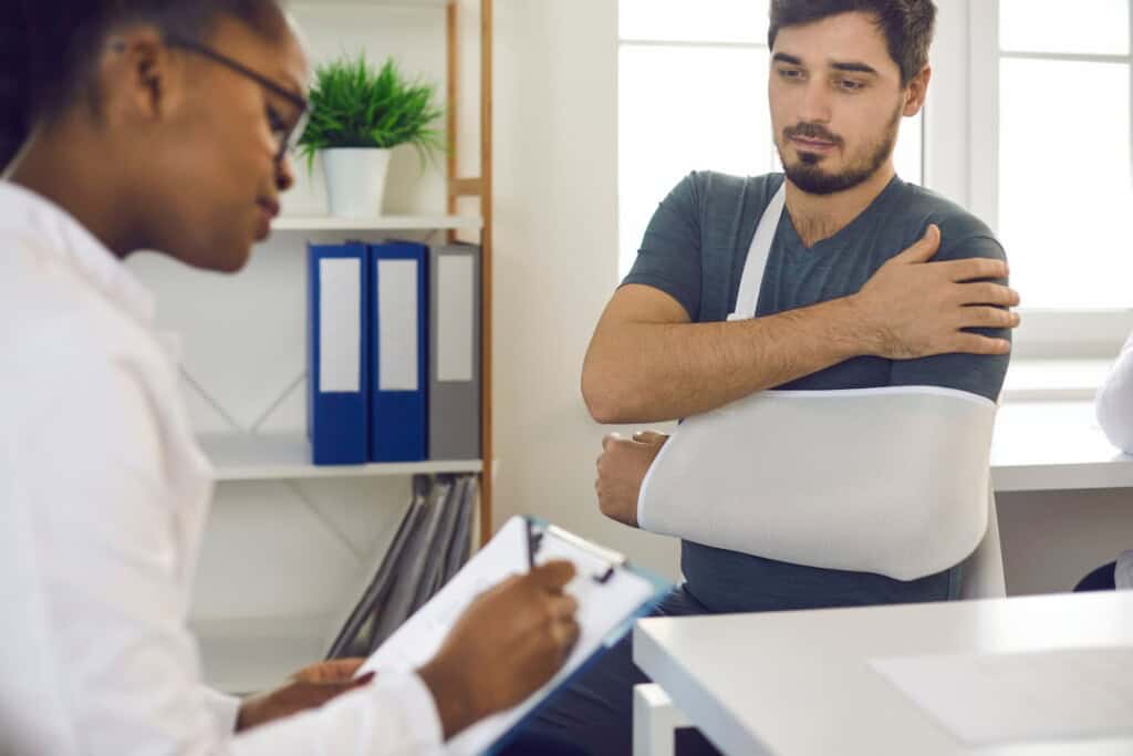 Female African-American doctor at hospital prescribing treatment and talking to young male patient wearing injury support sling.