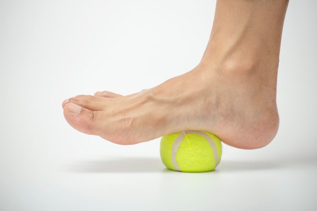 Tennis ball roll massage for plantar fascia stretch where a foot is pressing down on a yellow tennis ball against a white background.