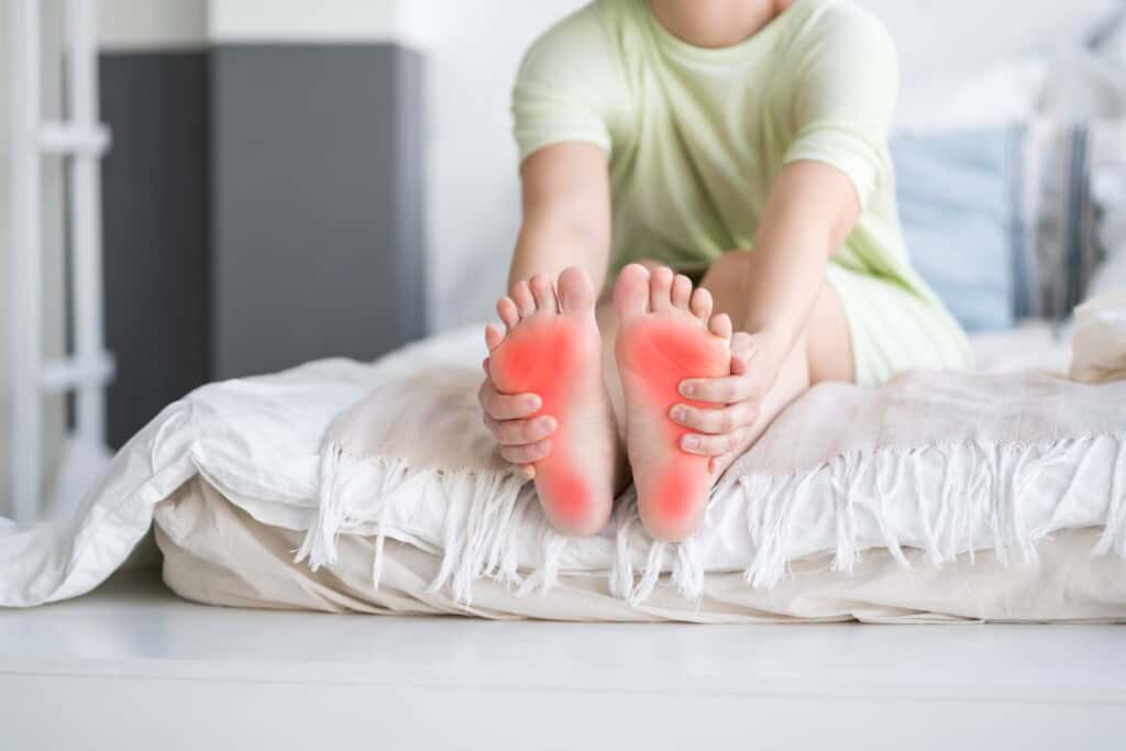 A woman sitting on a bed holding the bottom of her feet, which show red areas indicating pain caused plantar fasciitis.