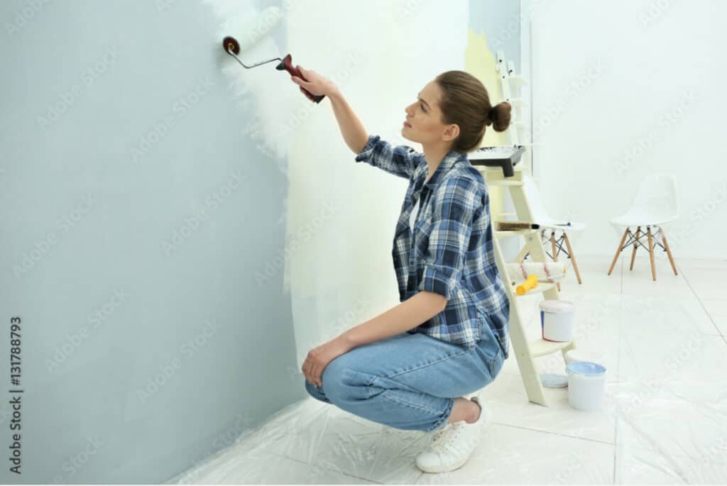 Beautiful young woman wearing jeans and a flannel shirt painting wall in room.