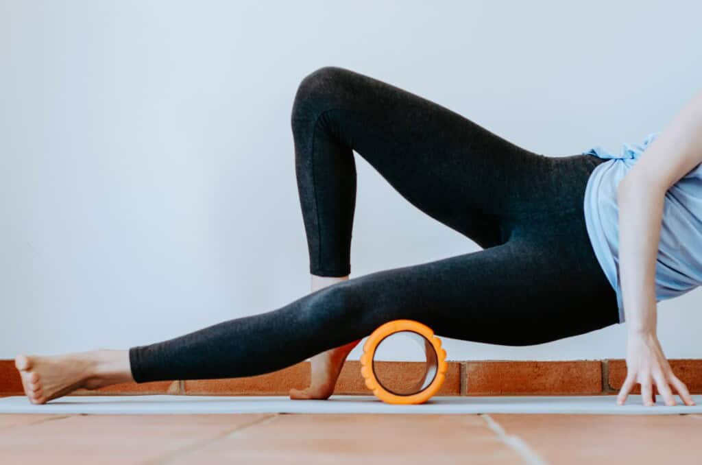 Woman using an orange foam roller on her thigh to alleviate IT Band Syndrome while performing a stretching exercise on a yoga mat.