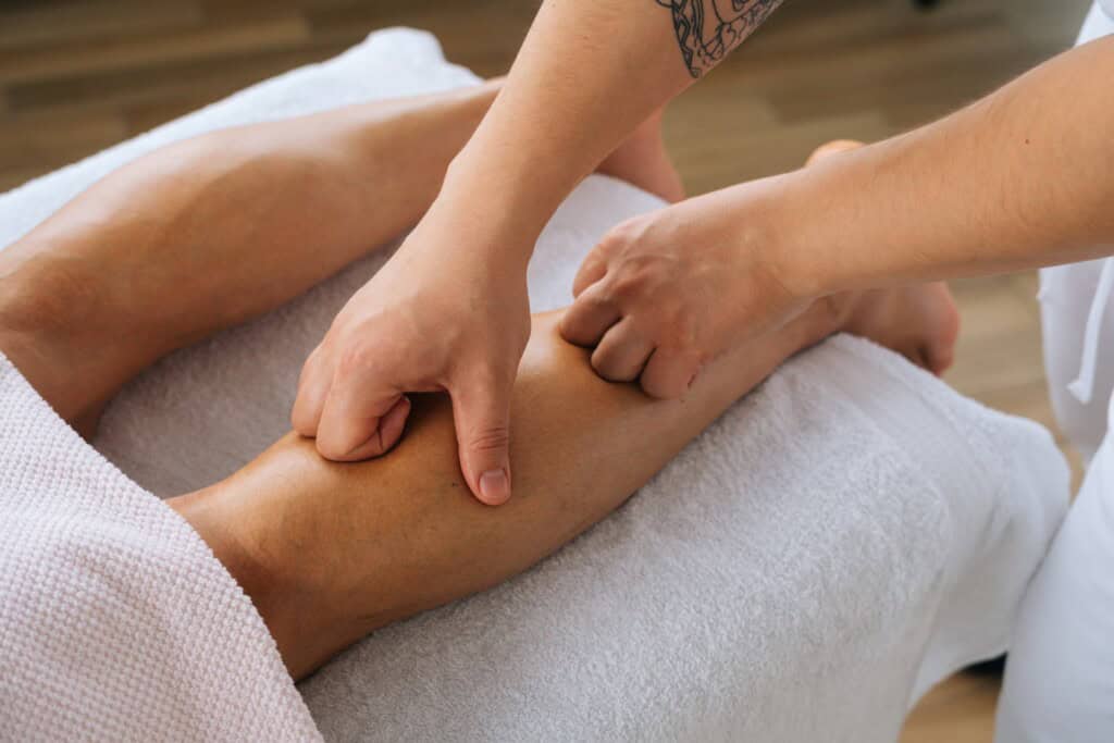 Close-up of a person's hands performing a leg massage on another person lying on a treatment table covered with a white towel, focusing on alleviating shin splints.