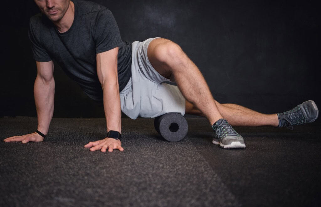 A man in a gray shirt and shorts uses a foam roller on the floor to stretch his right leg, targeting hip pain, while supporting himself with both hands on the ground.