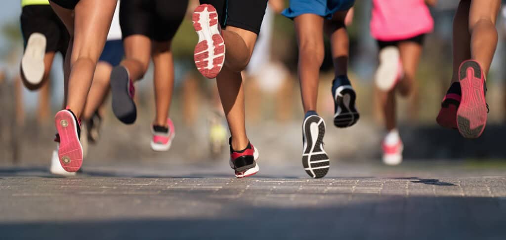 A group of runners in athletic shoes jogging on a paved path, with the focus on their lower legs and feet.