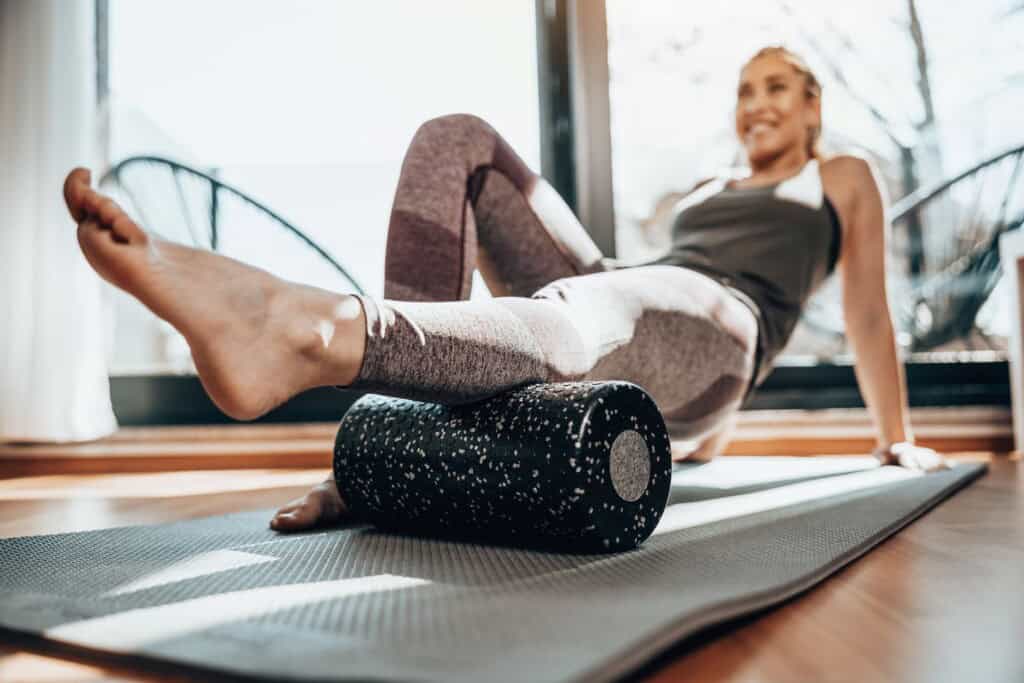 A woman in athletic wear uses a foam roller on a yoga mat indoors, addressing her shin splints, with natural light filtering through large windows.