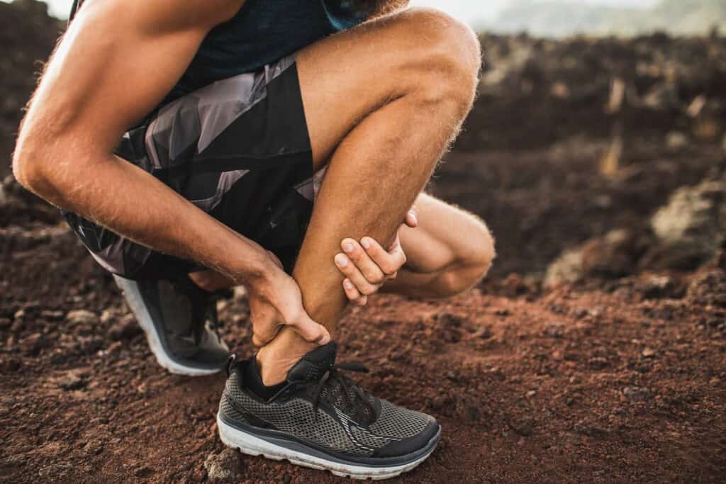 A close-up of a man's knees as he kneels on a running path, gripping his Achilles tendon in pain.