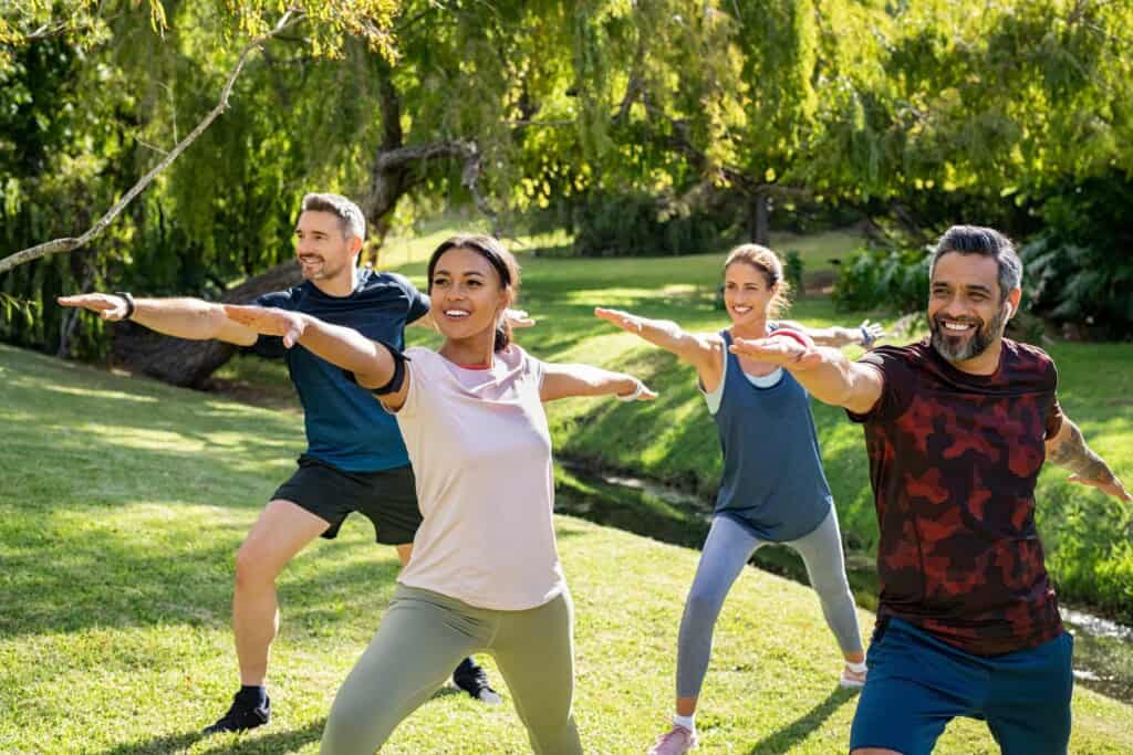 Group of four men and women in a park happily holding a warrior 2 yoga pose.