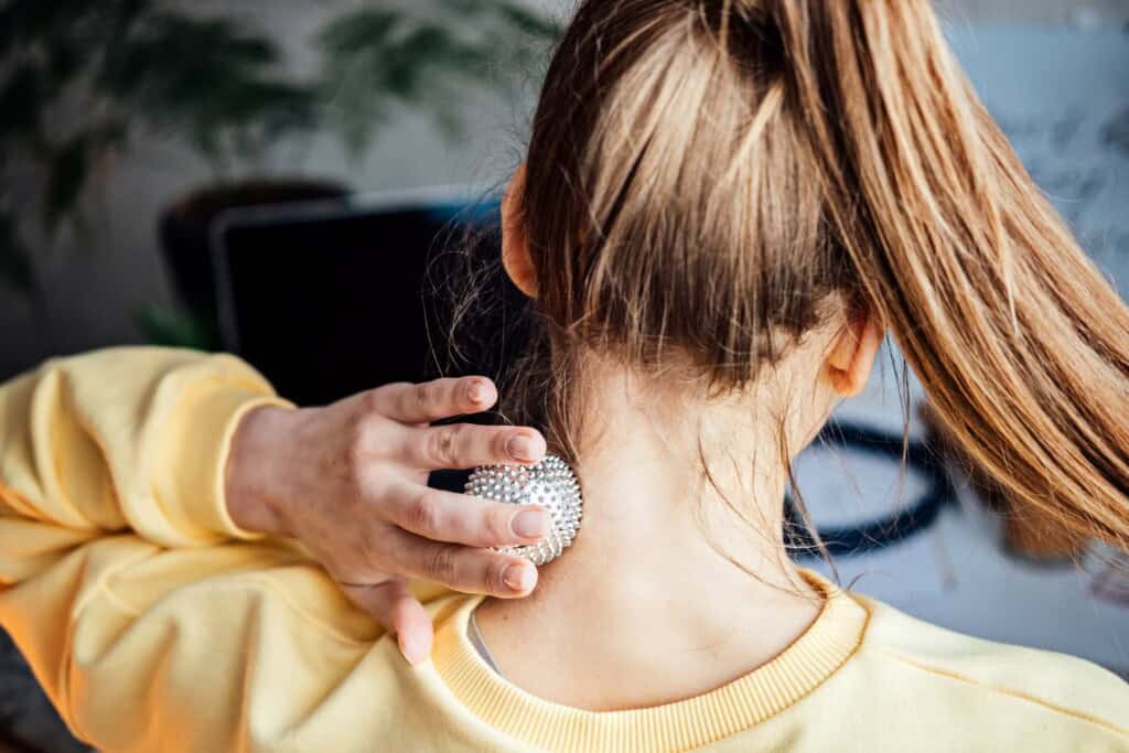 A woman with long hair in a ponytail wearing a yellow sweatshirt uses a spiky massage ball on the back of her neck, helping to relieve tension from forward head posture.