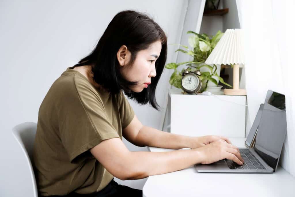 Asian woman sitting at a white desk, displaying forward head posture as she types on a laptop. A clock and a potted plant are on the desk near a window with a white curtain.