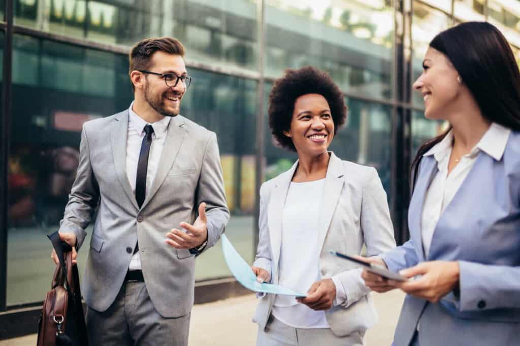 Three people in business attire are walking and smiling outside an office building. One holds a briefcase, another holds papers, and the third holds a tablet.