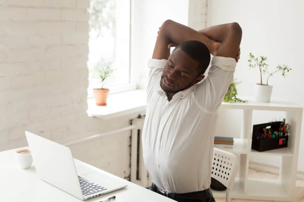 Tired African American man wearing a white shirt, sitting at a desk and stretching with his arms overhead beside an open laptop, relieving muscle tension after long hours of sitting.