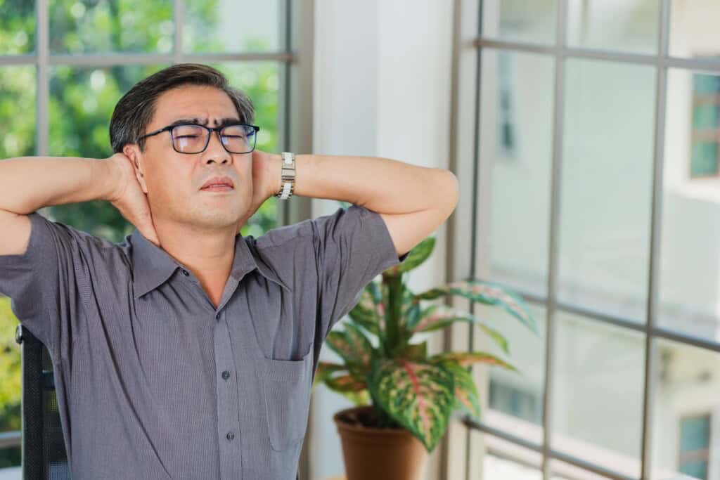 Asian senior businessman sitting at a desk, holding his neck with both hands, elbows out, and wincing in pain from prolonged sitting.