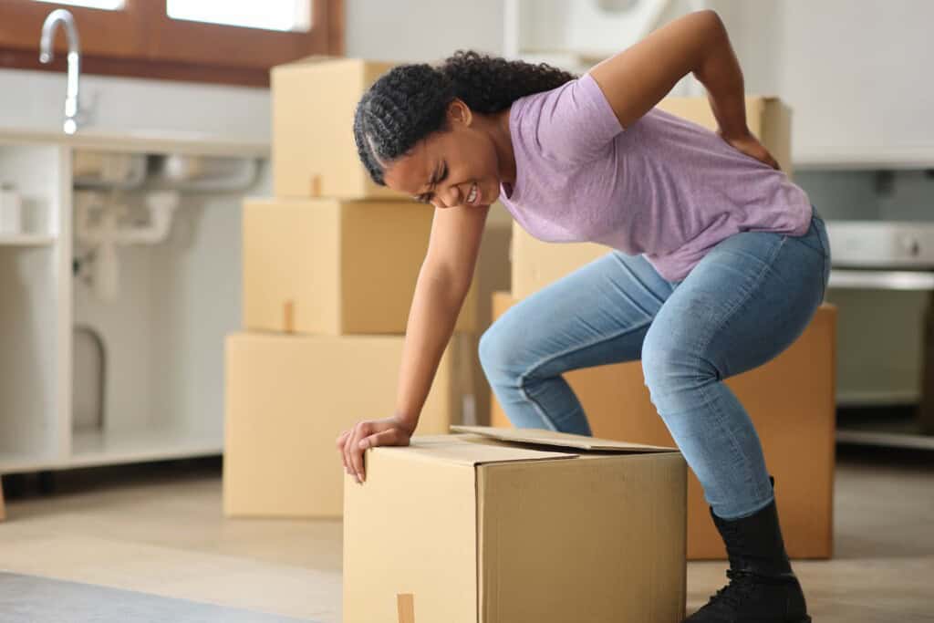 A woman bends over in pain while lifting a box, with moving boxes visible in the background.