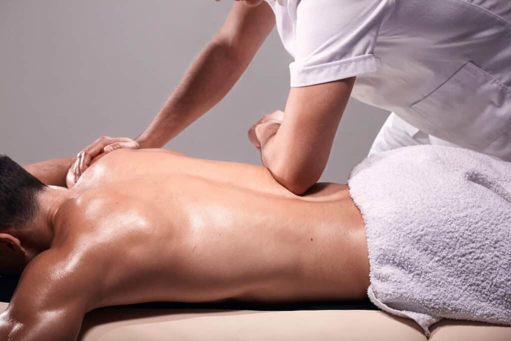 A person is receiving a back massage to alleviate back pain, from a therapist dressed in white, with a towel covering the lower body.