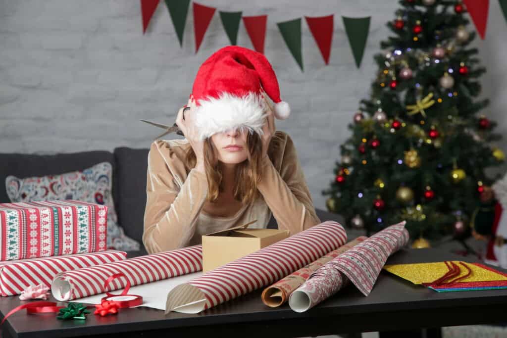 A stressed woman with a Santa hat pulled over her eyes, covering her ears with her hands at a gift-wrapping table surrounded by festive holiday decor.