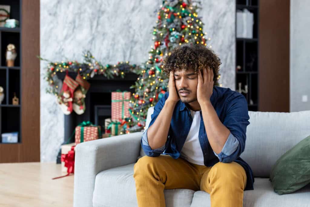 African American man holding his head in pain, sitting on a sofa near a decorated Christmas tree, showing signs of holiday stress.