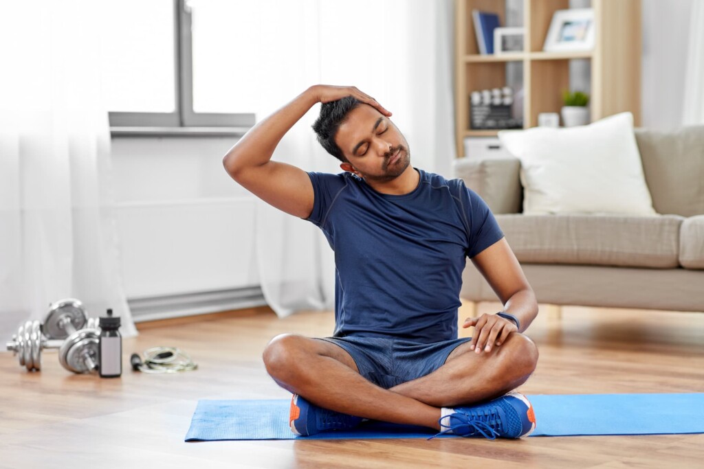 A man sits cross-legged on a yoga mat, stretching his neck with one hand.