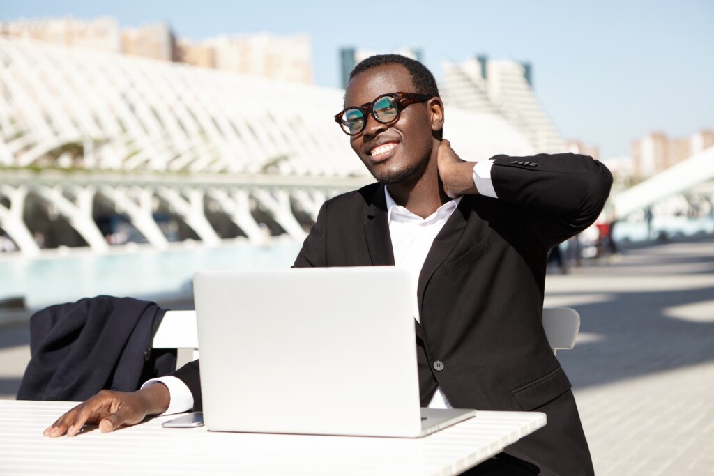 A person in a black suit sits at an outdoor table with a laptop, smiling comfortably and touching their neck, relieved after recovering from whiplash. The background shows modern architecture against a clear sky.