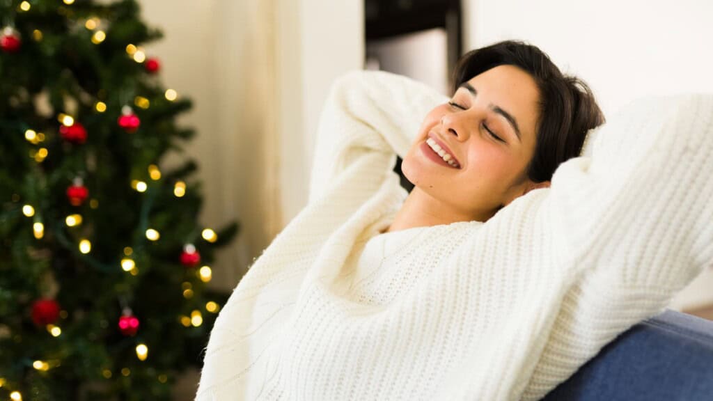 A smiling woman stretches with her arms overhead at her desk, in front of a laptop and papers, surrounded by festive decorations. 