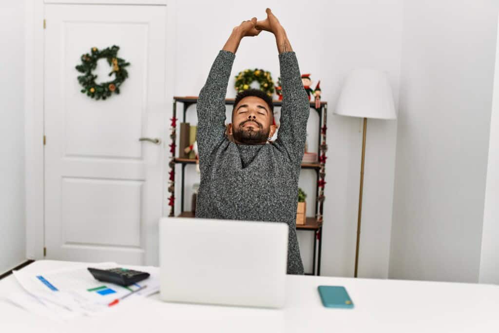 A man stretching with hisarms overhead at a desk, surrounded by festive decorations, laptop, and papers, finding a moment of relief from holiday stress.