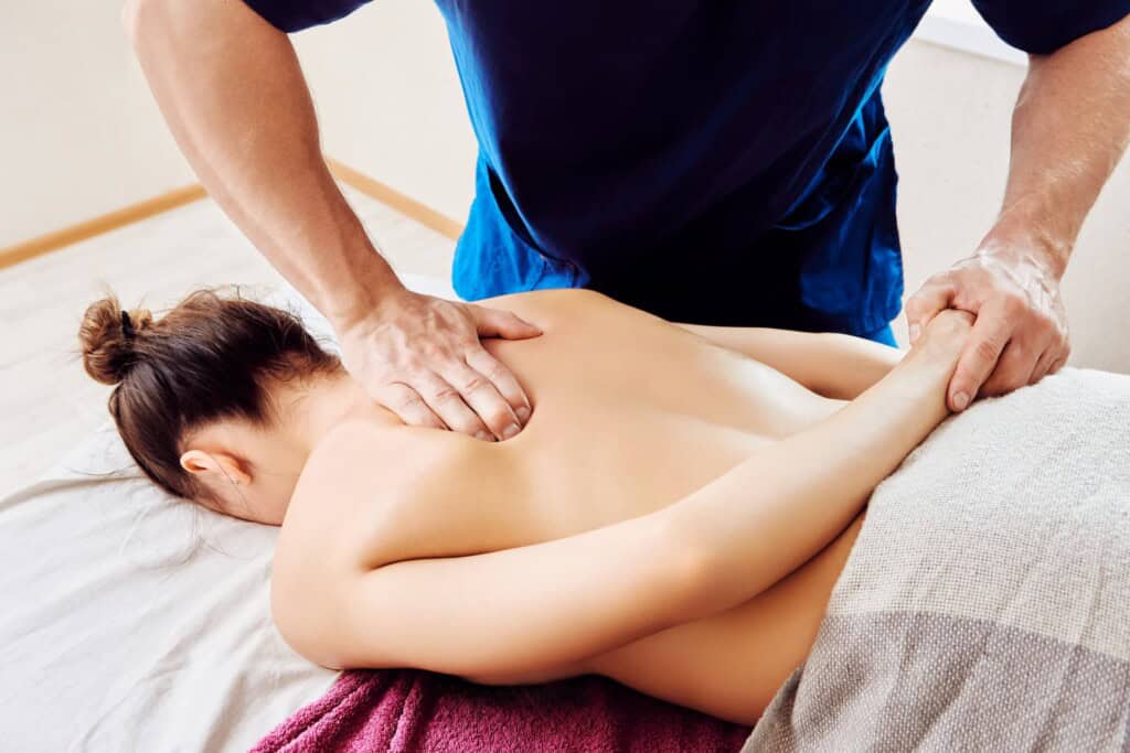 Bodyworker applying deep tissue pressure to a young woman's upper back near the shoulder blade.