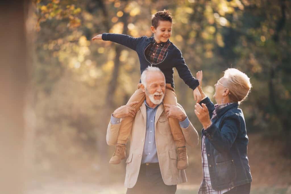 Grandparents enjoying a walk in a sunlit, wooded area with their grandson. The grandfather carries the smiling boy on his shoulders as the boy stretches his arms wide, while the grandmother walks beside them, also smiling.