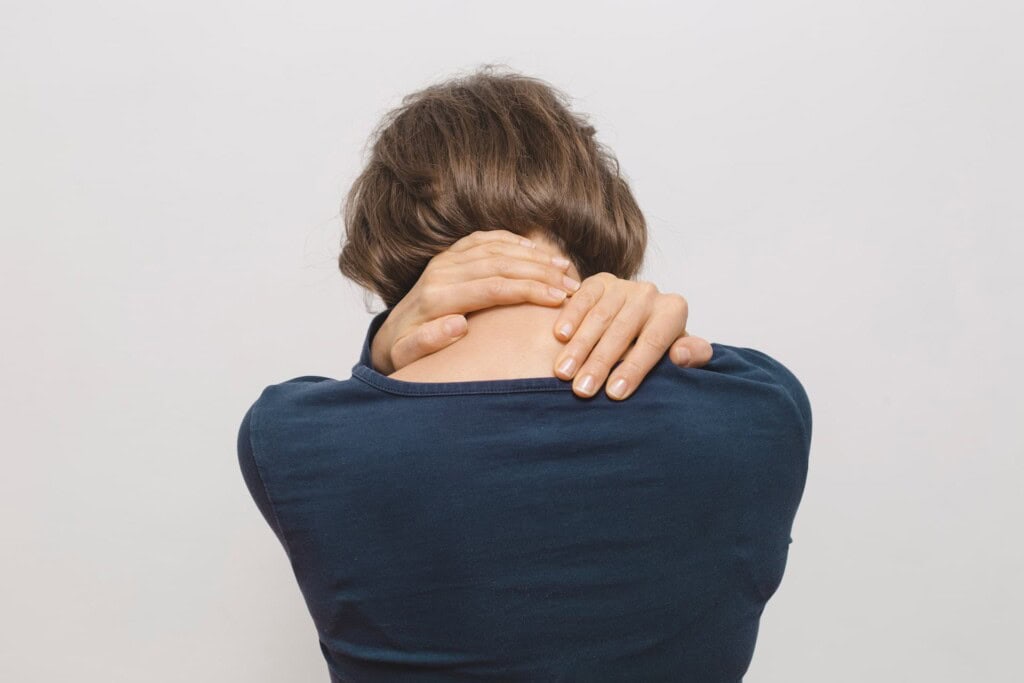 A woman in a blue shirt rubbing the back of her neck with both hands due to upper back pain.