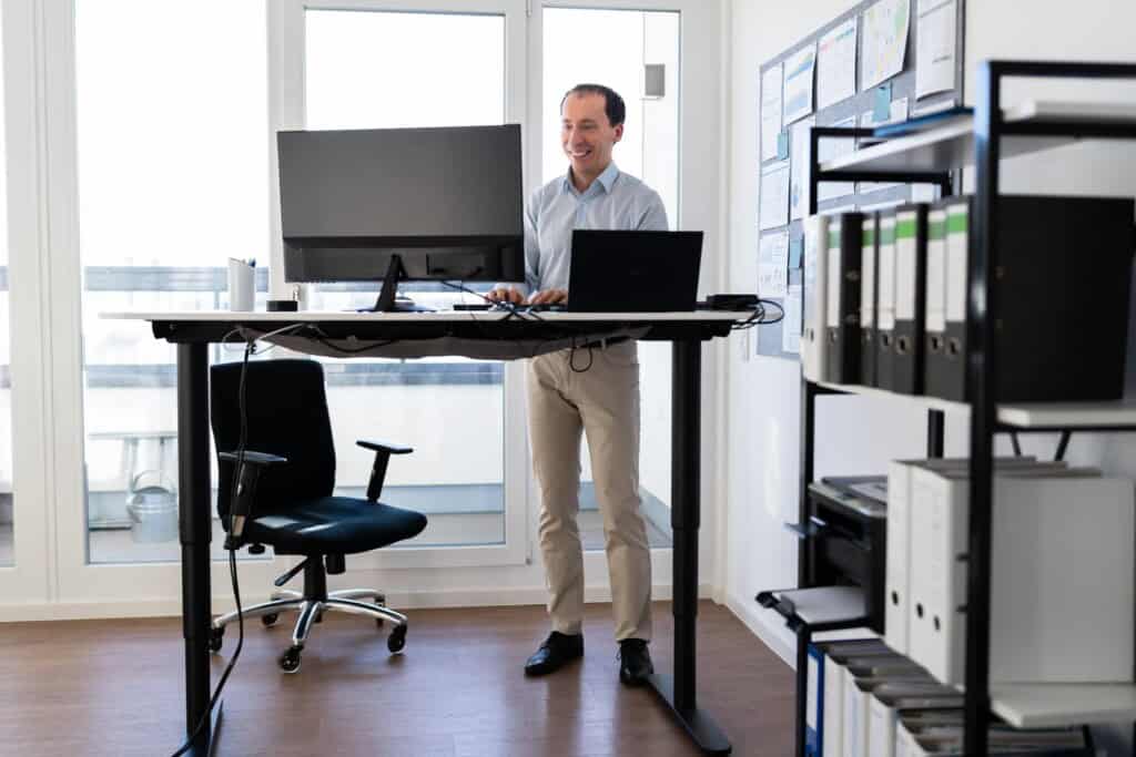 A man stands at a height-adjustable desk with a computer and laptop in a bright office, smiling. The ergonomic setup is ideal for alleviating back pain. Shelves with files are in the background.