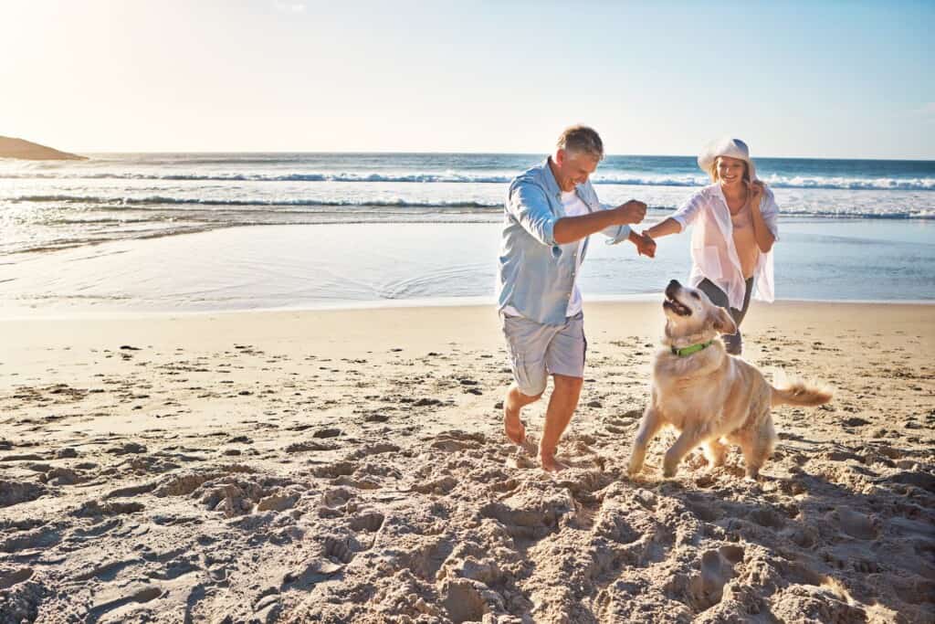 Happy older couple walks on the beach, holding hands and playing with their dog by the sea.